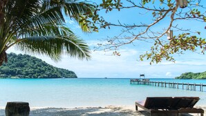 Ein Strand mit einer Holzbank und einem Stein, ein klares blaues Meer und ein Steg, der sich ins Wasser erstreckt.