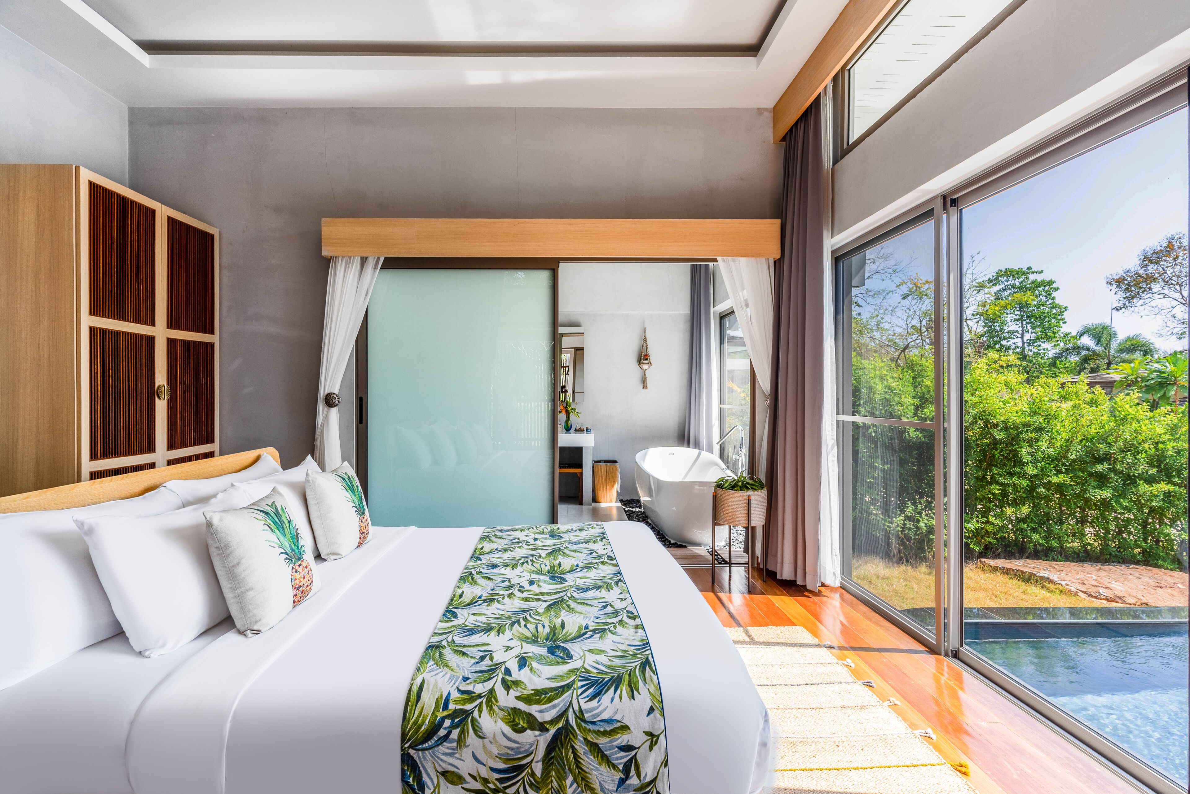 A modern bedroom with a large bed, a wooden wardrobe, a bathroom with a bathtub, and a view of a swimming pool and greenery.