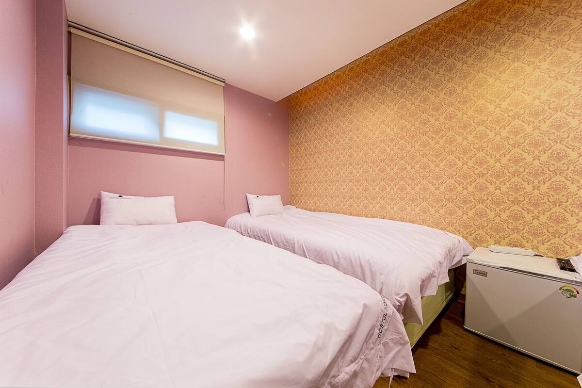 A bedroom with two beds, a small refrigerator, and a window with blinds.