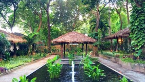 Ein friedlicher Garten mit einem zentralen Wasserspiel, umgeben von üppiger Vegetation und strohgedeckten Gebäuden.