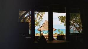Ein Zimmer mit Bett, einem Fenster mit Blick auf einen Strand und einem Gebäude mit Strohdach.