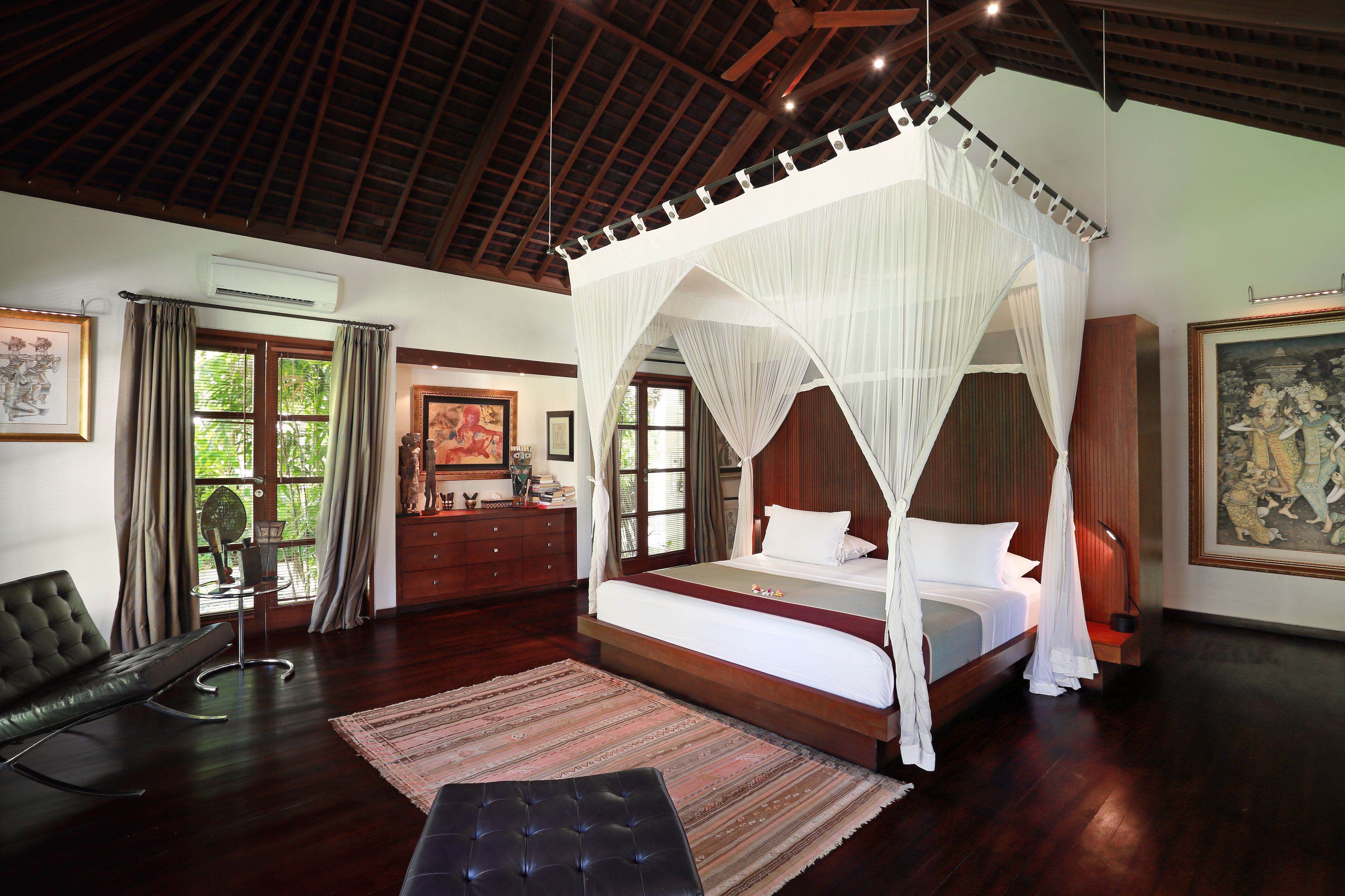A spacious bedroom with a four-poster bed, a sitting area with a sofa and armchair, and a large window offering a view of greenery.