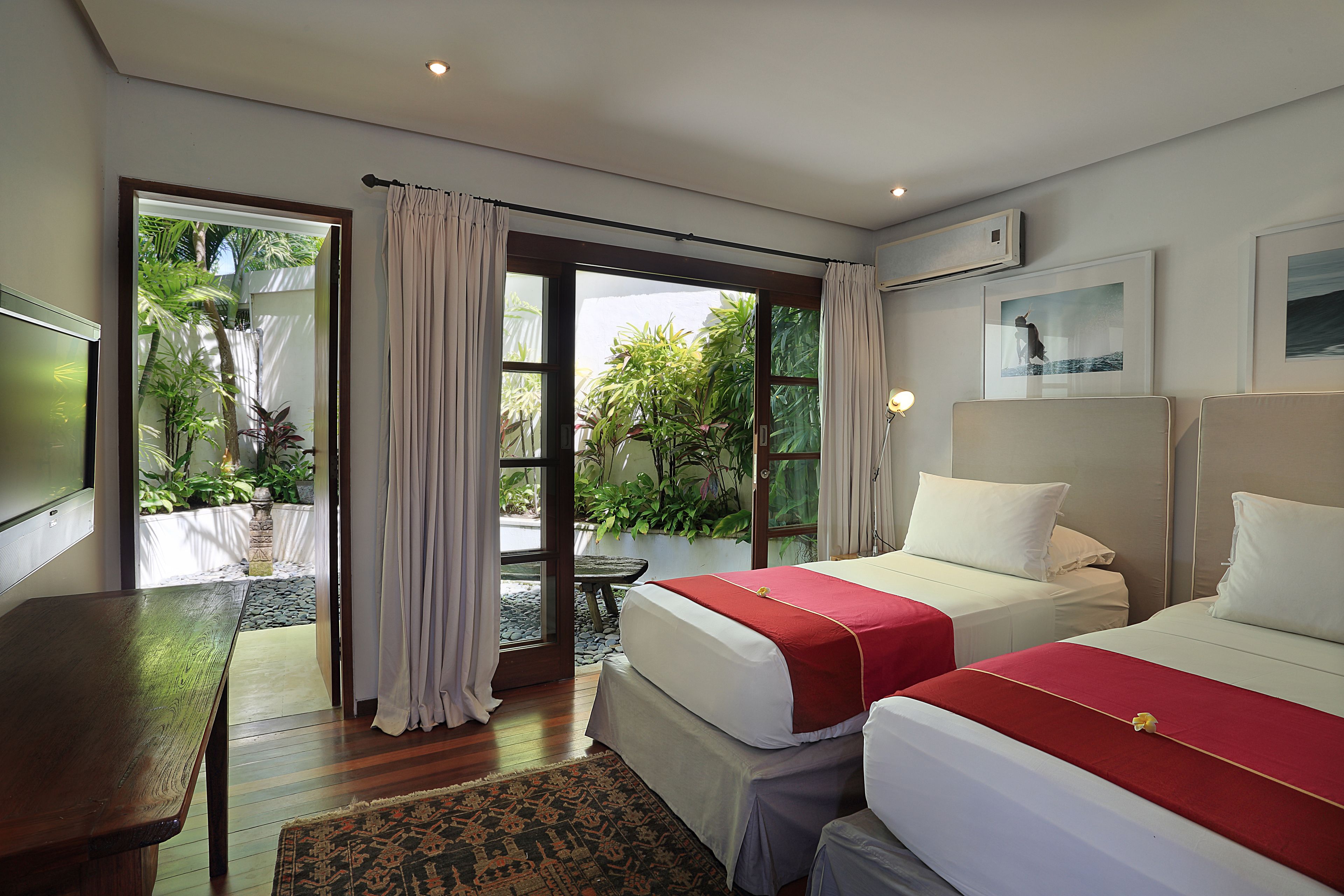 Habitación de hotel con dos camas, una mesa de madera y vista al jardín a través de la puerta corrediza de vidrio.