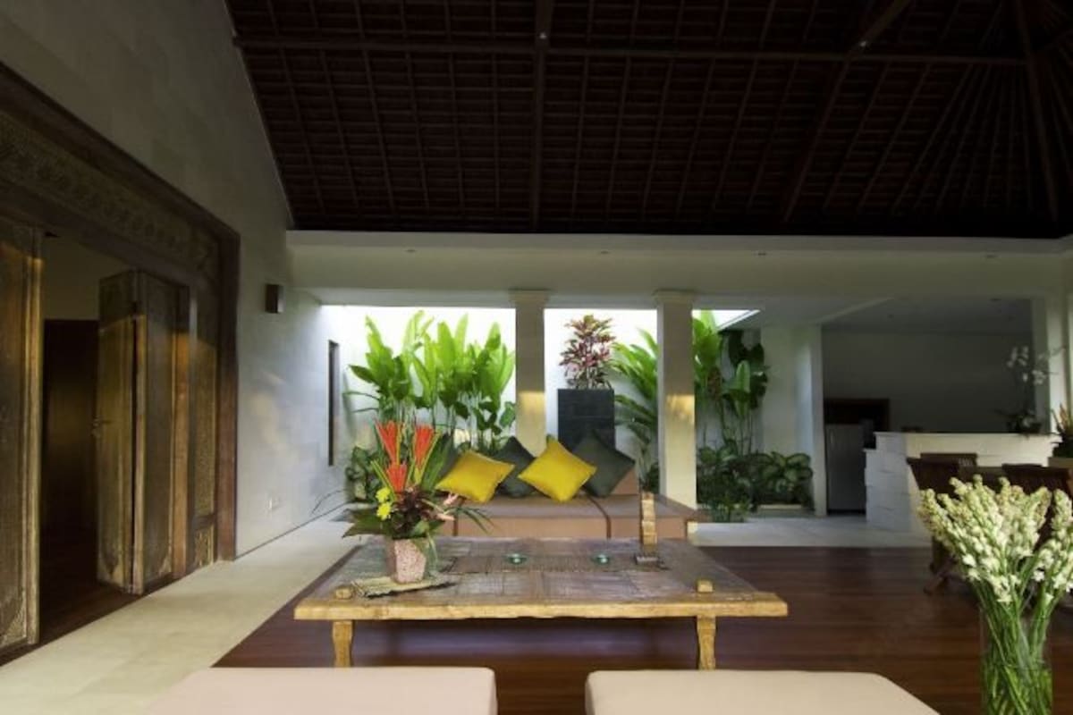Amplia sala de estar con una mesa de madera, asientos cómodos y vistas a un jardín exuberante a través de grandes ventanales.