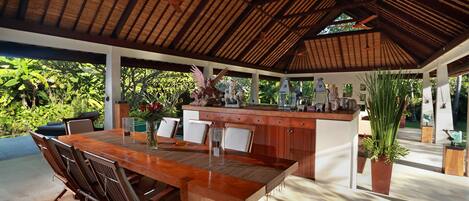 A covered outdoor dining area with a wooden table and chairs, surrounded by lush greenery.
