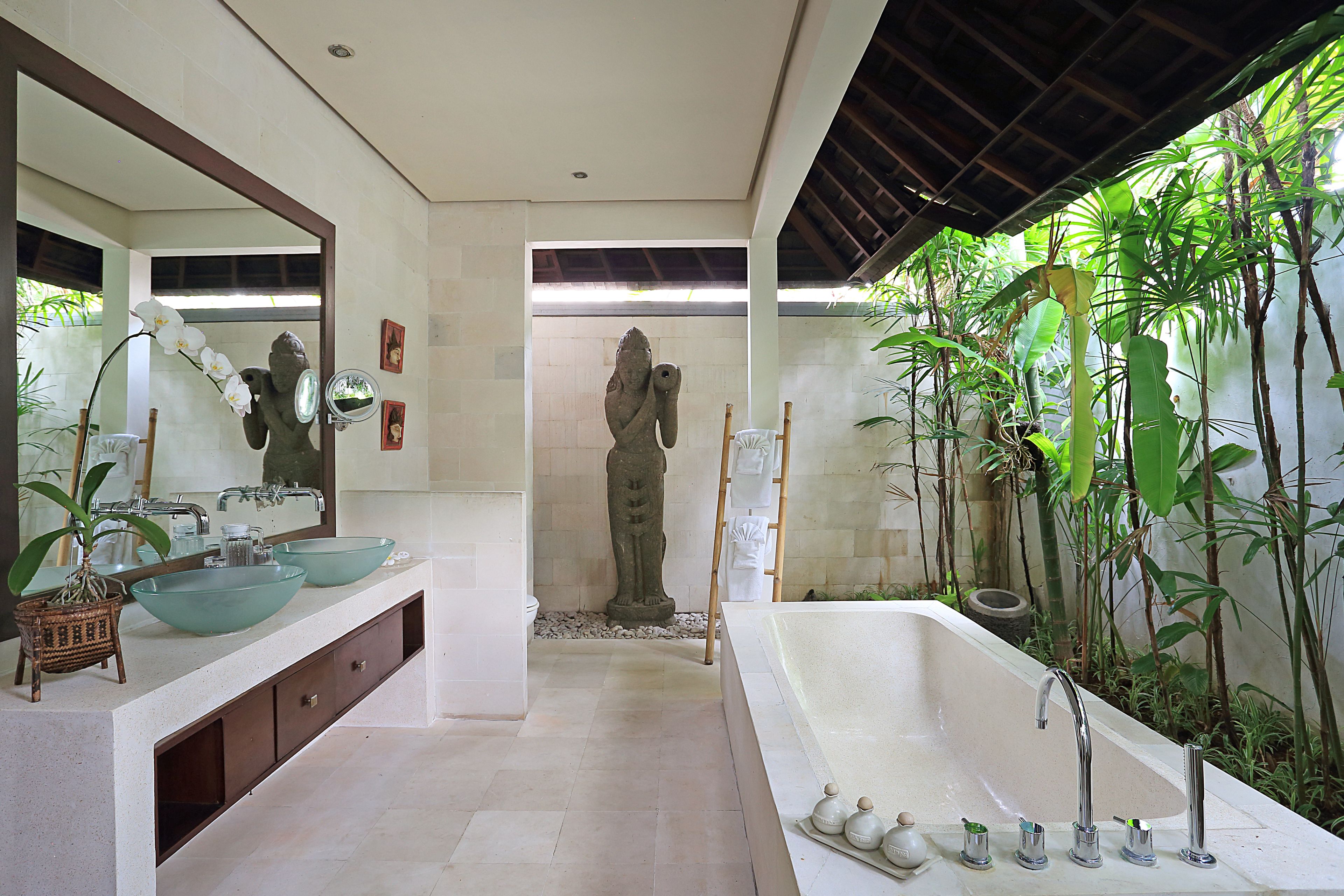 A modern bathroom with a large bathtub, two sinks with glass bowls, a stone statue, and lush greenery.