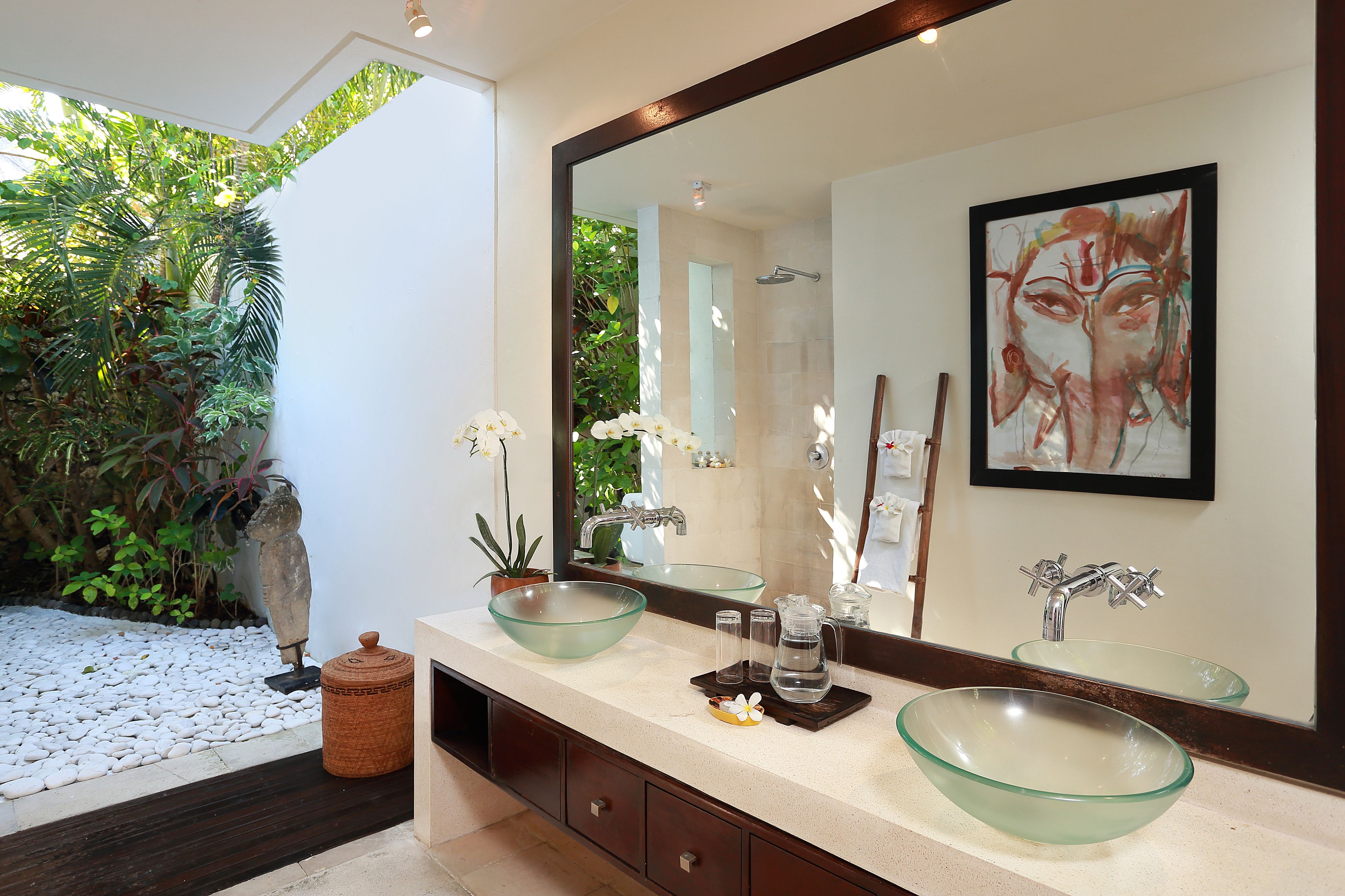 A bathroom with a large mirror, two glass bowls on the counter, a potted plant, and a shower area visible through the glass door.