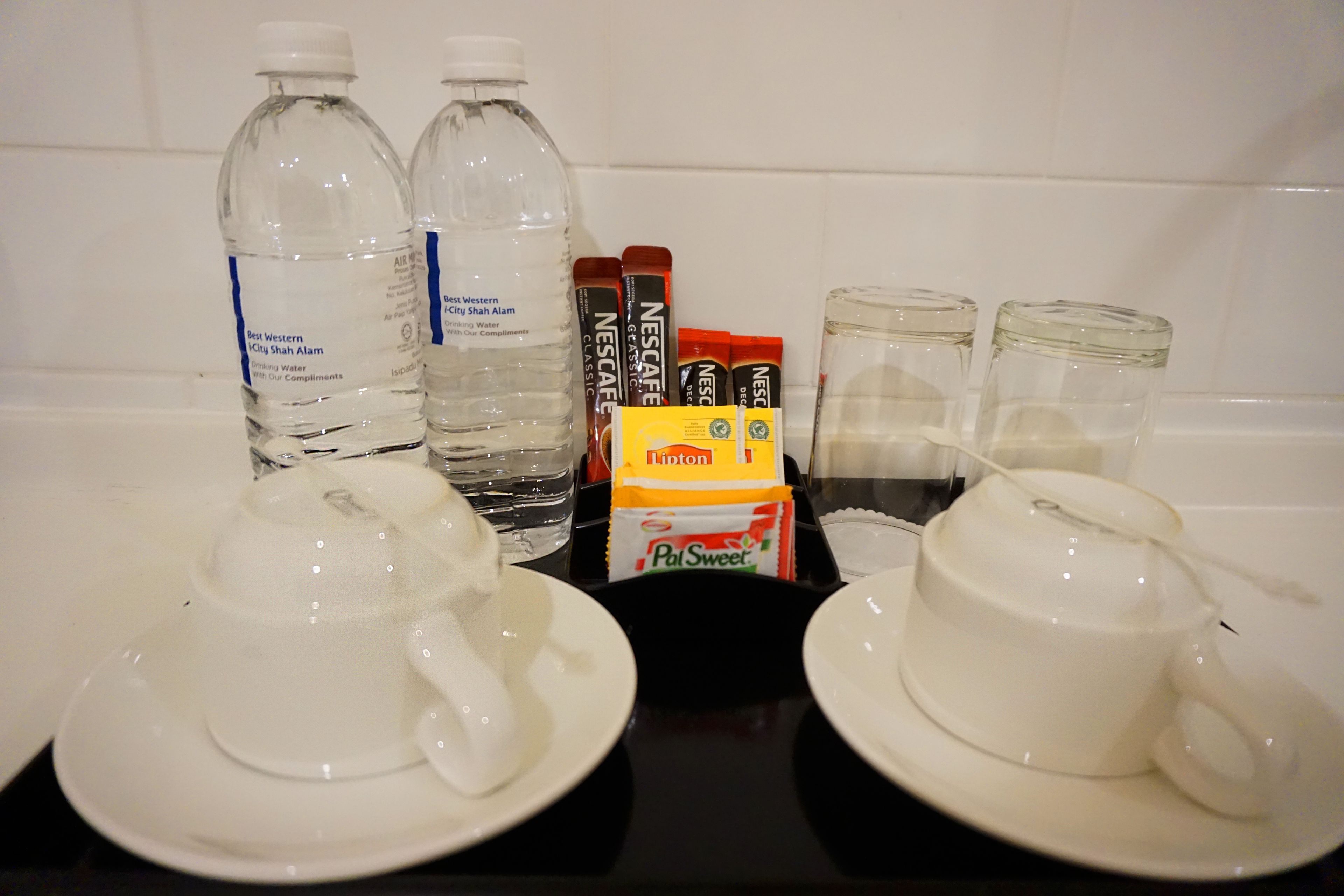 Une table avec deux bouteilles d’eau, deux sachets de café Nescafé, un sachet de sucre Pol Sweet et deux tasses vides sur des assiettes blanches.