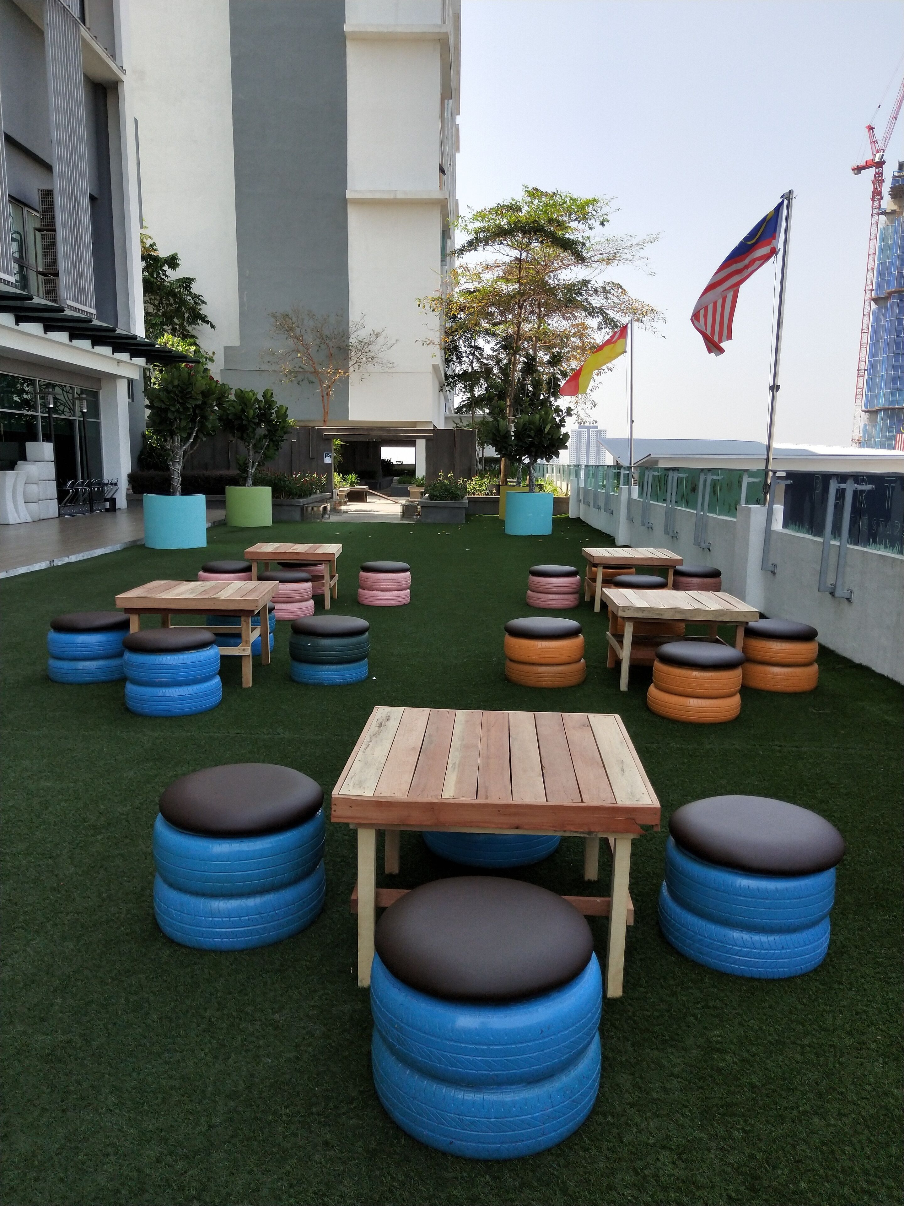 An outdoor patio with wooden tables and blue and orange tire stools, surrounded by artificial grass.