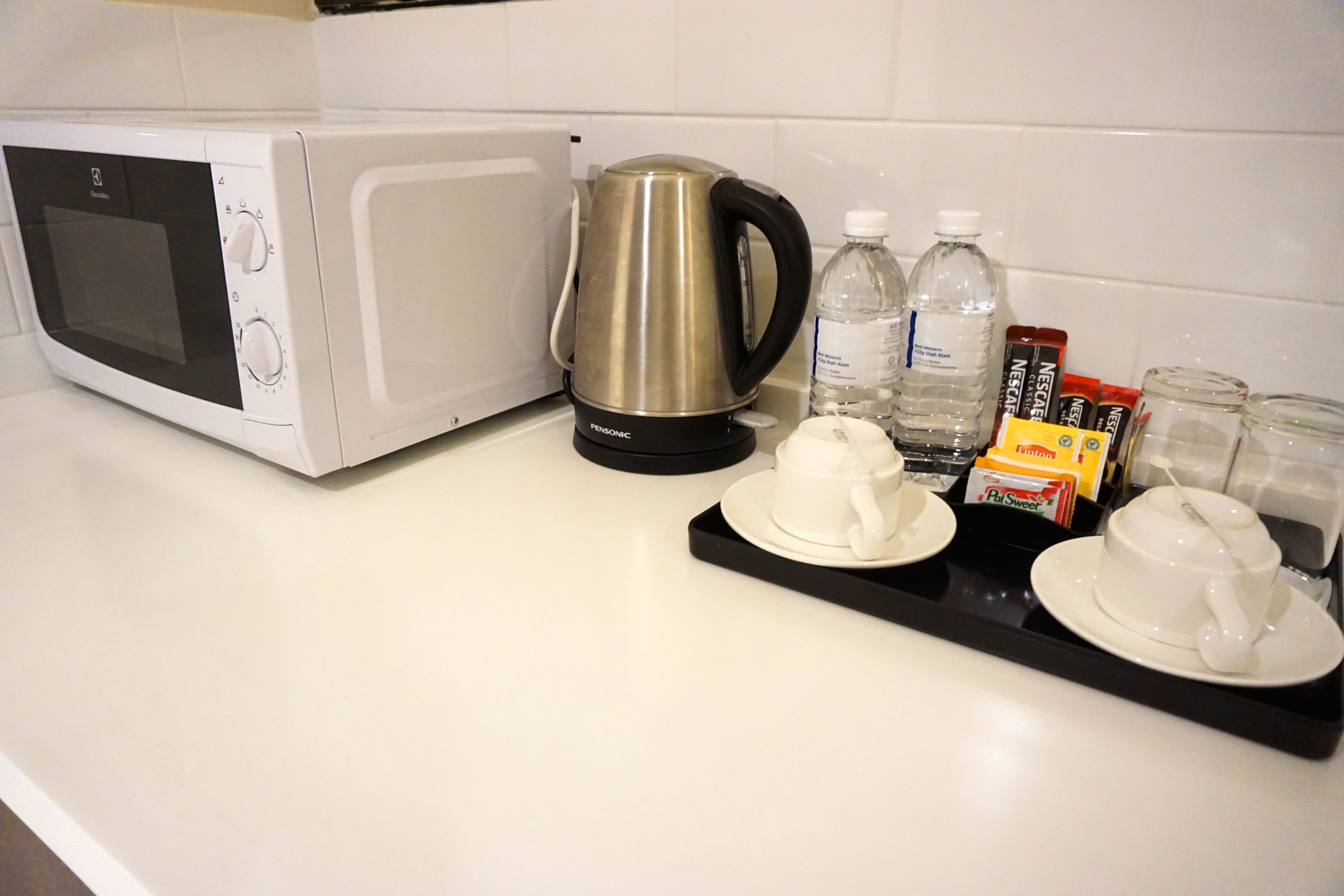 A kitchen counter with a white microwave, a black coffee maker, water bottles, and a tray with cups and a box of instant coffee.