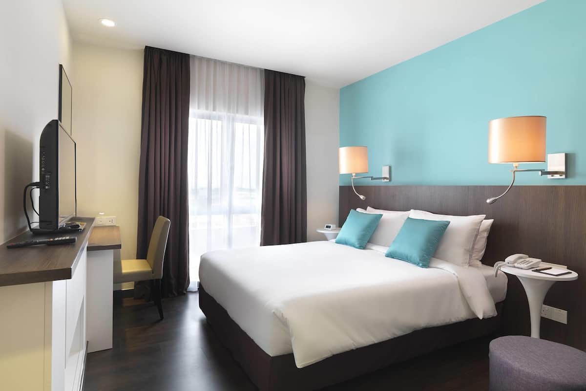 A hotel room with a large bed, a desk with a computer, a chair, a small table with a phone, and two wall-mounted lamps.