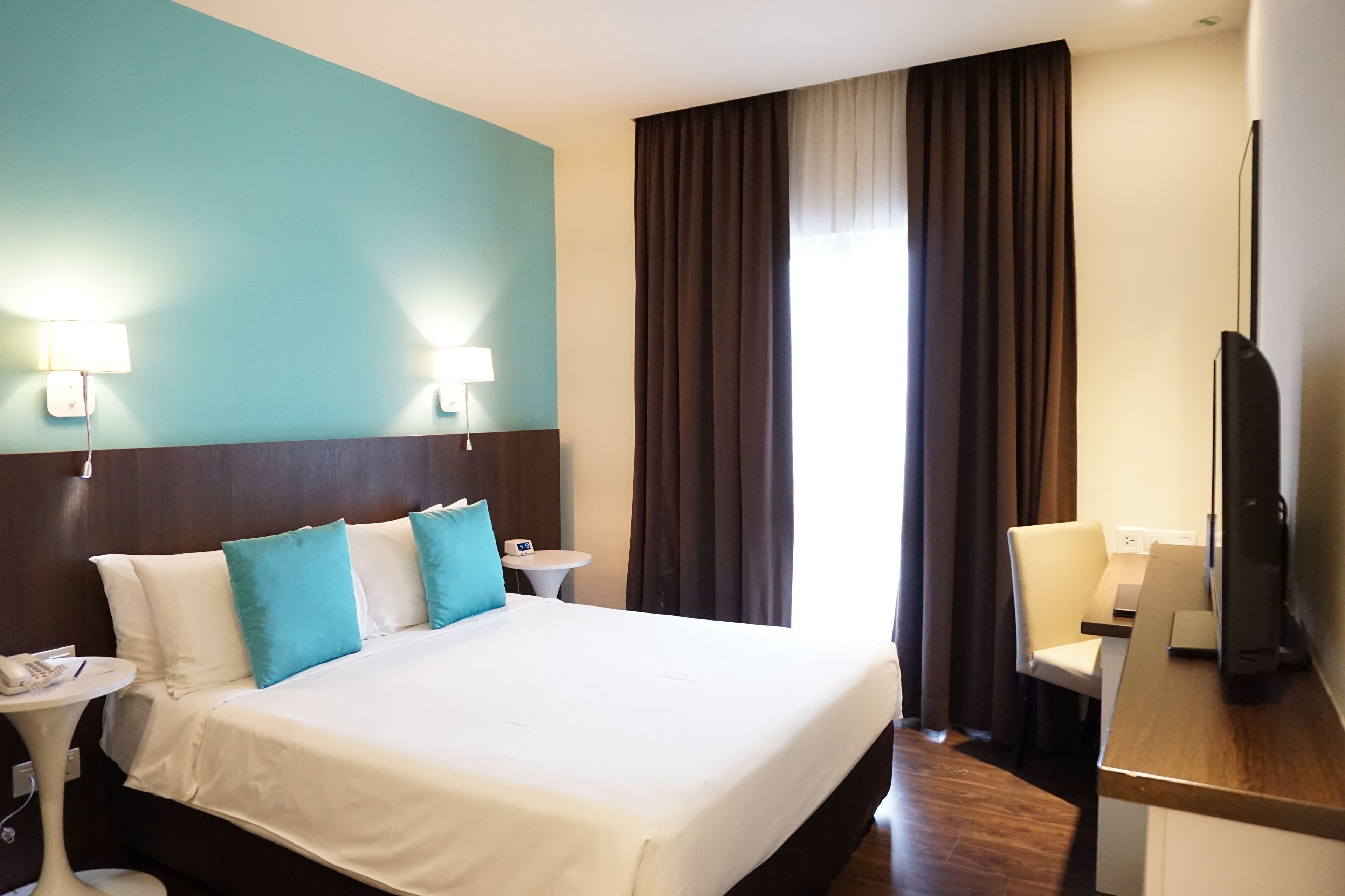 A hotel room with a large bed, two bedside lamps, a desk with a chair, a TV, and a window with curtains.