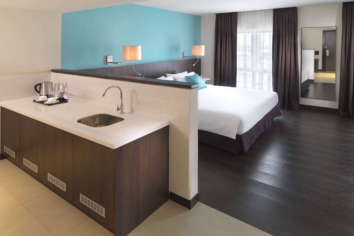 A hotel room with a kitchenette, a bed, a large mirror, and a bathroom.