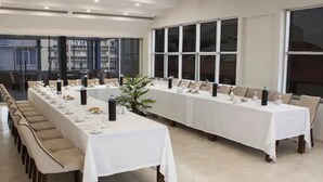 Una larga mesa de conferencias con sillas, manteles blancos y botellas de agua, ubicada en una sala con ventanas amplias y piso de baldosas.