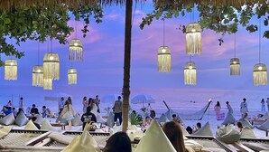 Un área de descanso frente a la playa con lámparas colgantes, techo de paja y sillones reclinables con cojines.