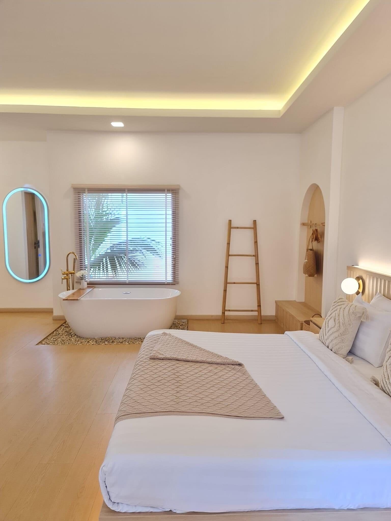 A modern bedroom with a large bed, a bathtub, a window with blinds, a ladder, and a mirror with a light.