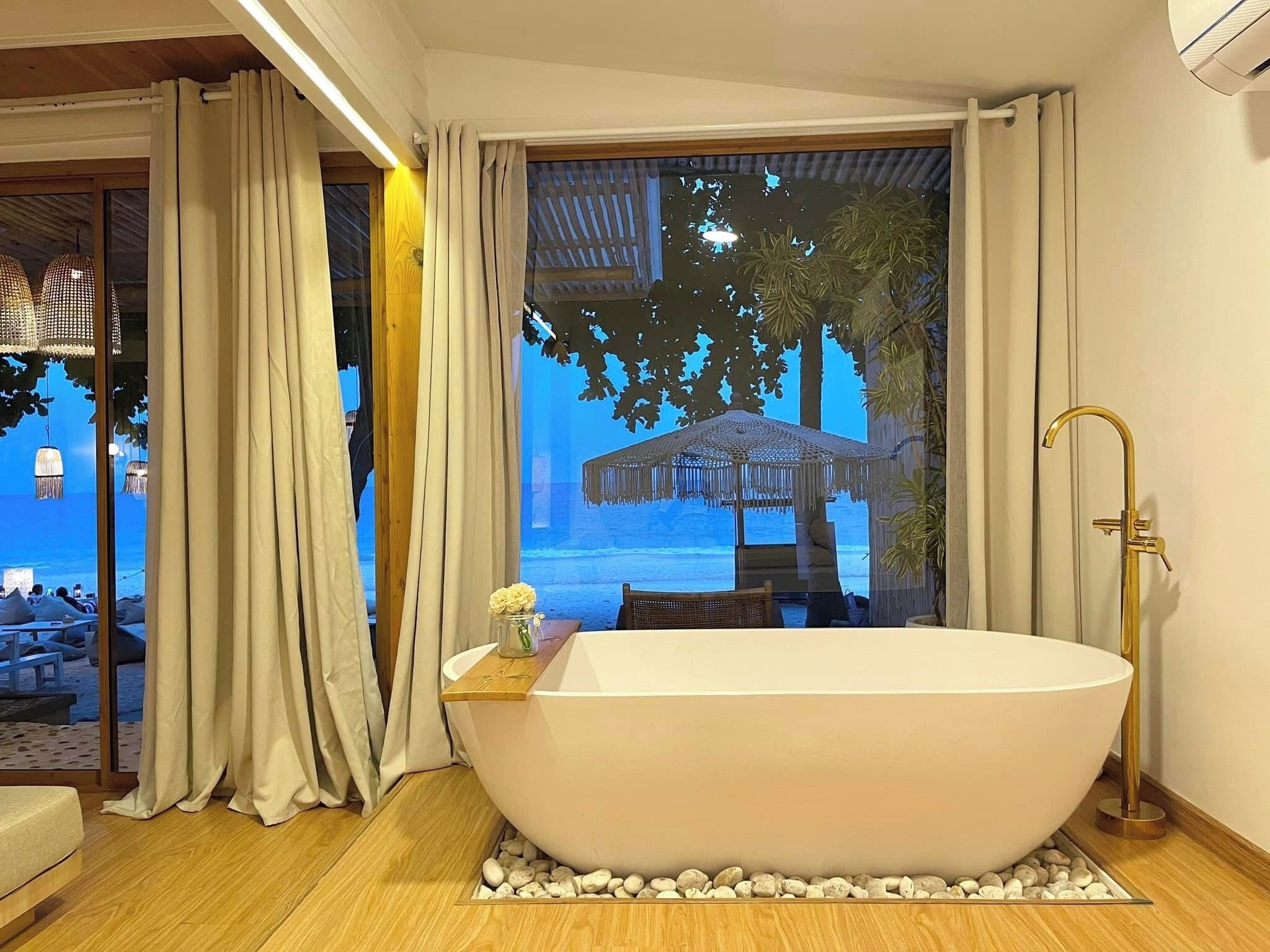 A modern bathroom with a freestanding bathtub, a gold faucet, and a view of a beach through a large window.