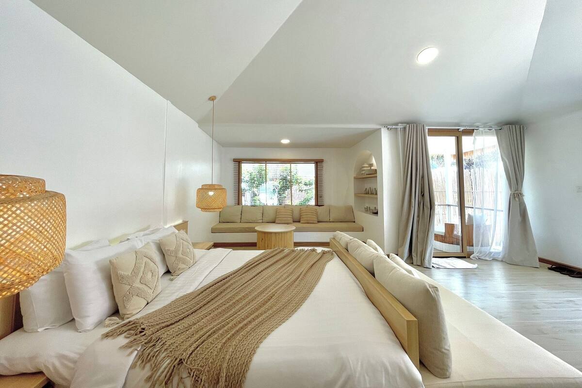 Quarto com cama grande, sofá, mesa pequena e janela com cortinas.
