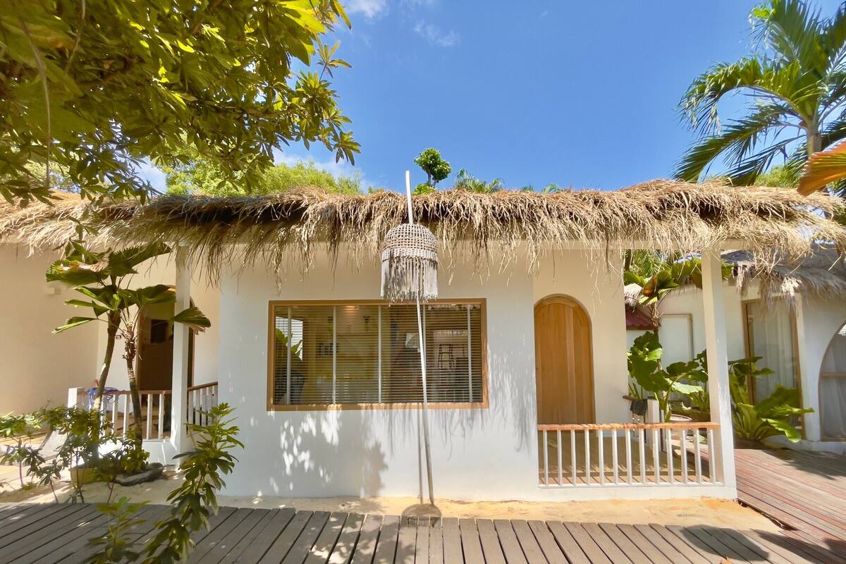 Uma cabana com telhado de palha, varanda de madeira, janela com persianas e luminária pendente.