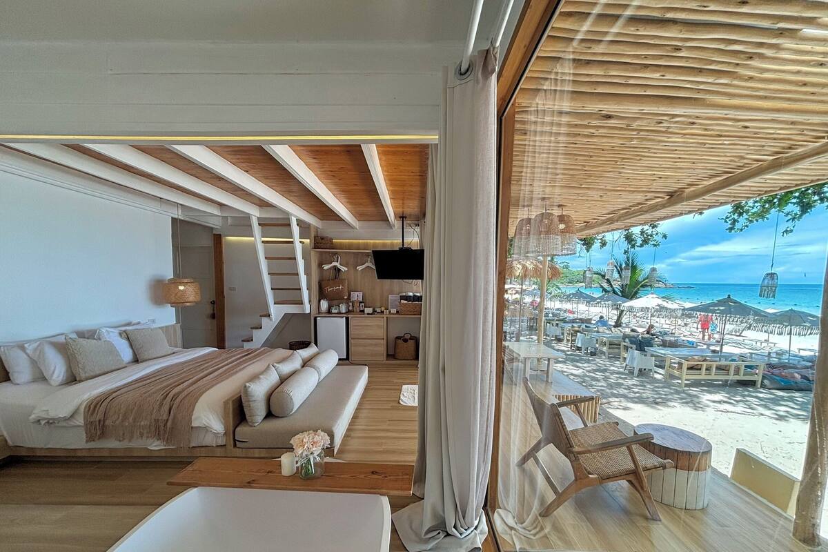 Quarto moderno com cama grande, sofá, cozinha compacta e vista para a praia.
