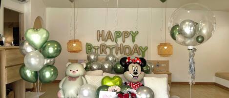 Una habitación con una cama, una mesa redonda de madera, globos y un cartel que dice 'HAPPY BIRTHDAY'.