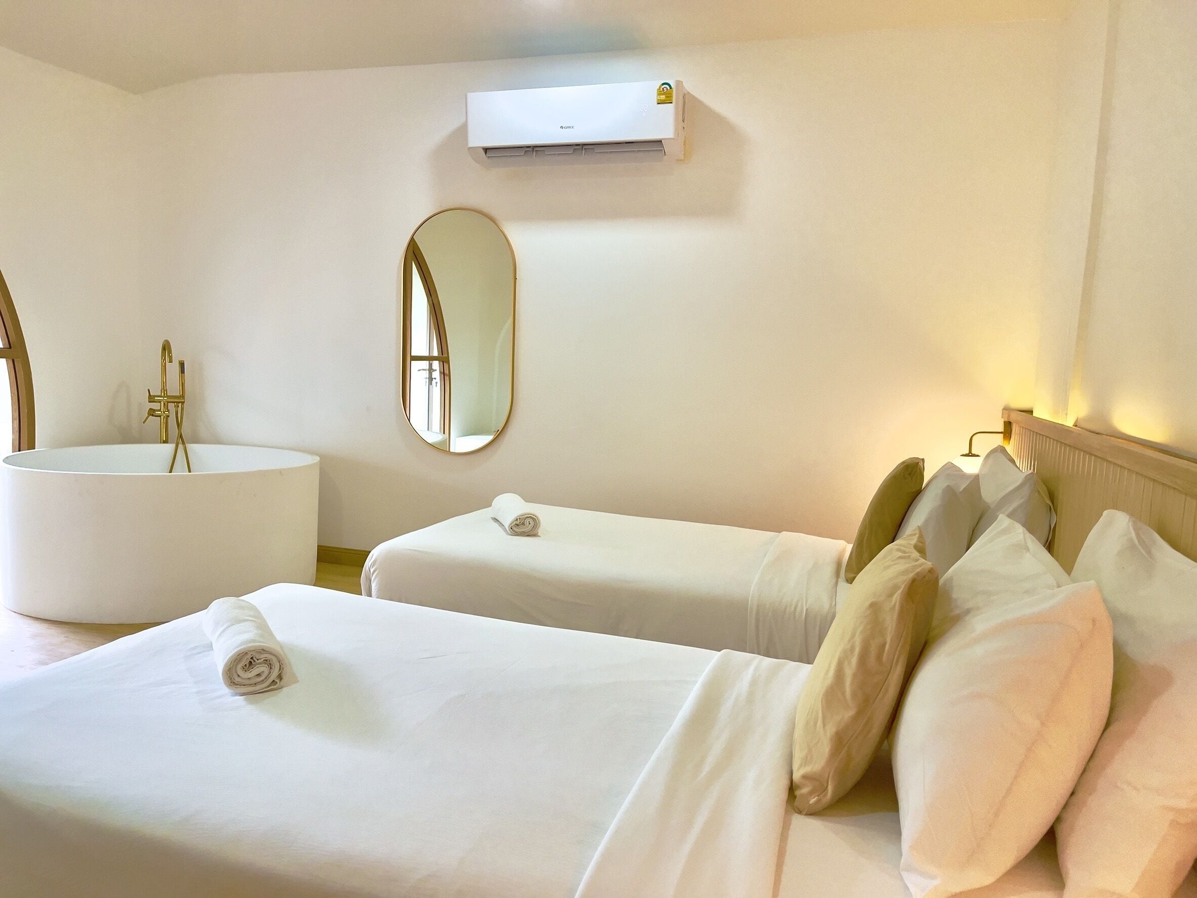 Una habitación de hotel con un lavamanos grande y blanco, dos camas con sábanas blancas, un espejo redondo y un equipo de aire acondicionado en la pared.