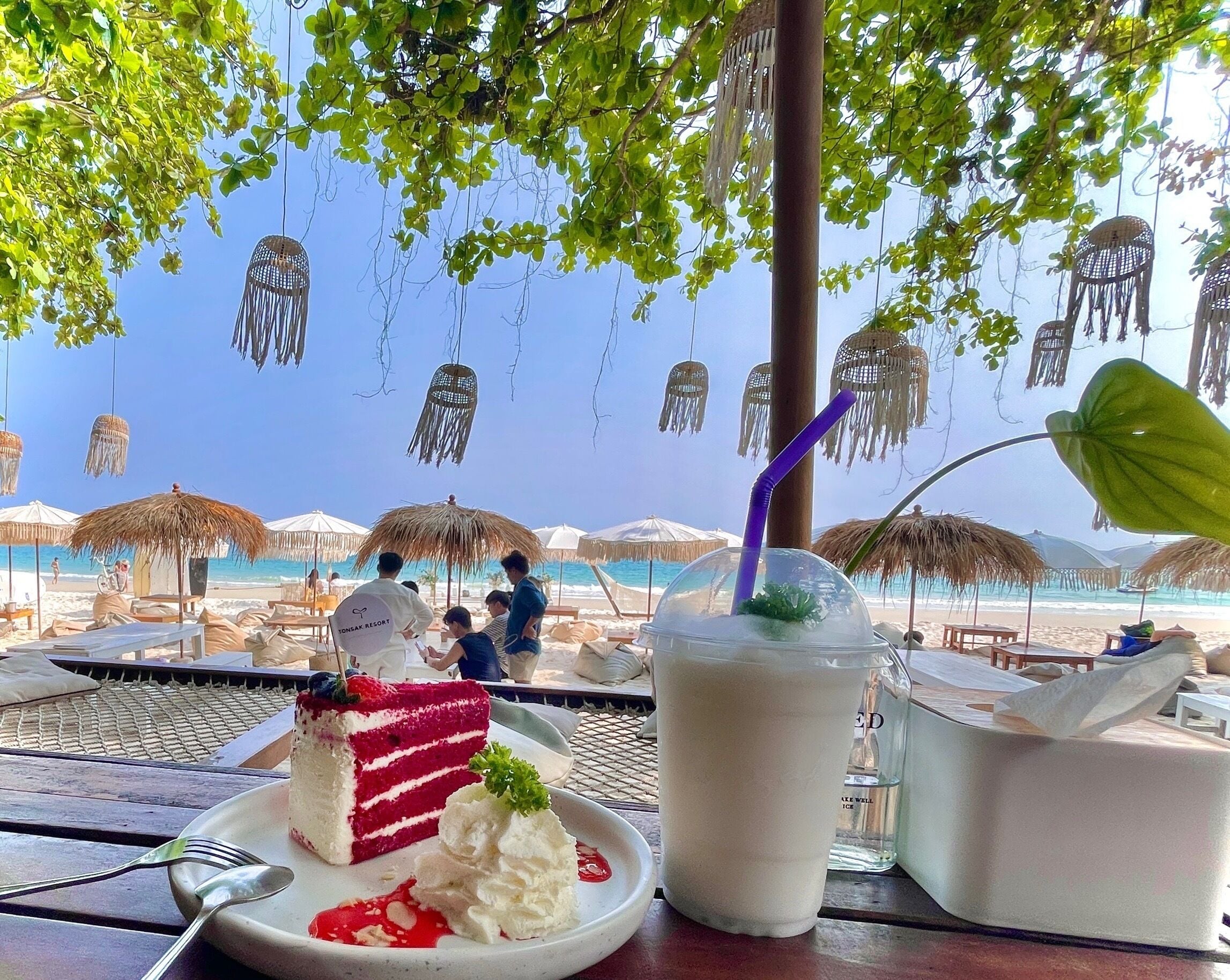 Un espace de restauration aménagé sur la plage, avec des parasols en chaume, une table où sont disposés un dessert et une boisson, et une vue sur l’océan.