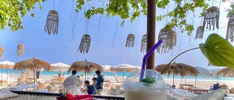 Un espace de restauration aménagé sur la plage, avec des parasols en chaume, une table où sont disposés un dessert et une boisson, et une vue sur l’océan.