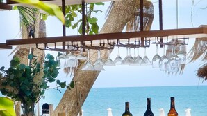 Un bar frente a la playa con mostrador de madera, copas de vino colgantes y botellas de vino.