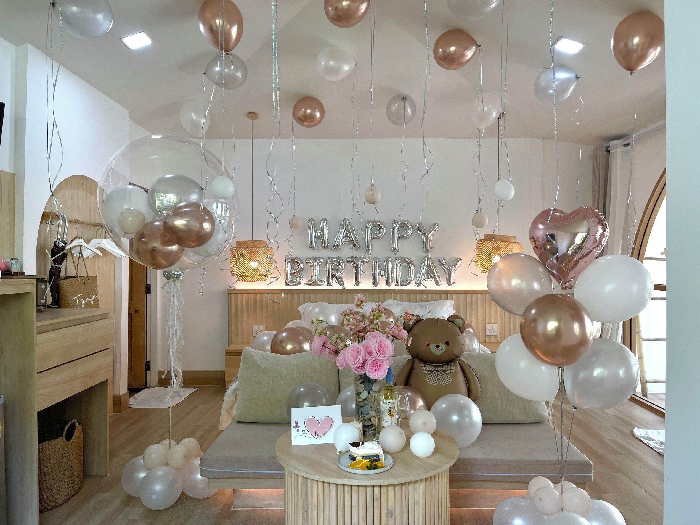 A room with a sofa, a table with a cake and a card, balloons, and a teddy bear.
