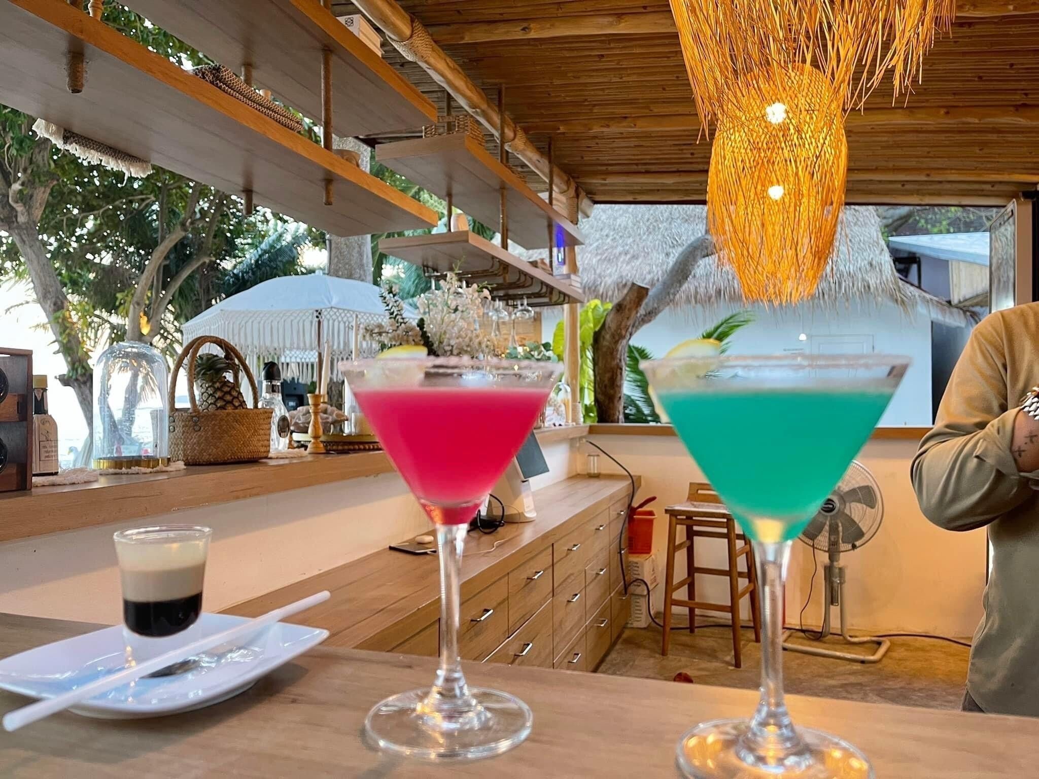Angolo bar con due cocktail su un bancone di legno, una ventola e una persona sullo sfondo.