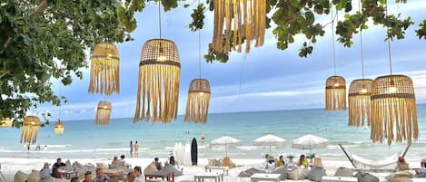 A beachside restaurant with outdoor seating, hanging wicker lamps, and a view of the sea.