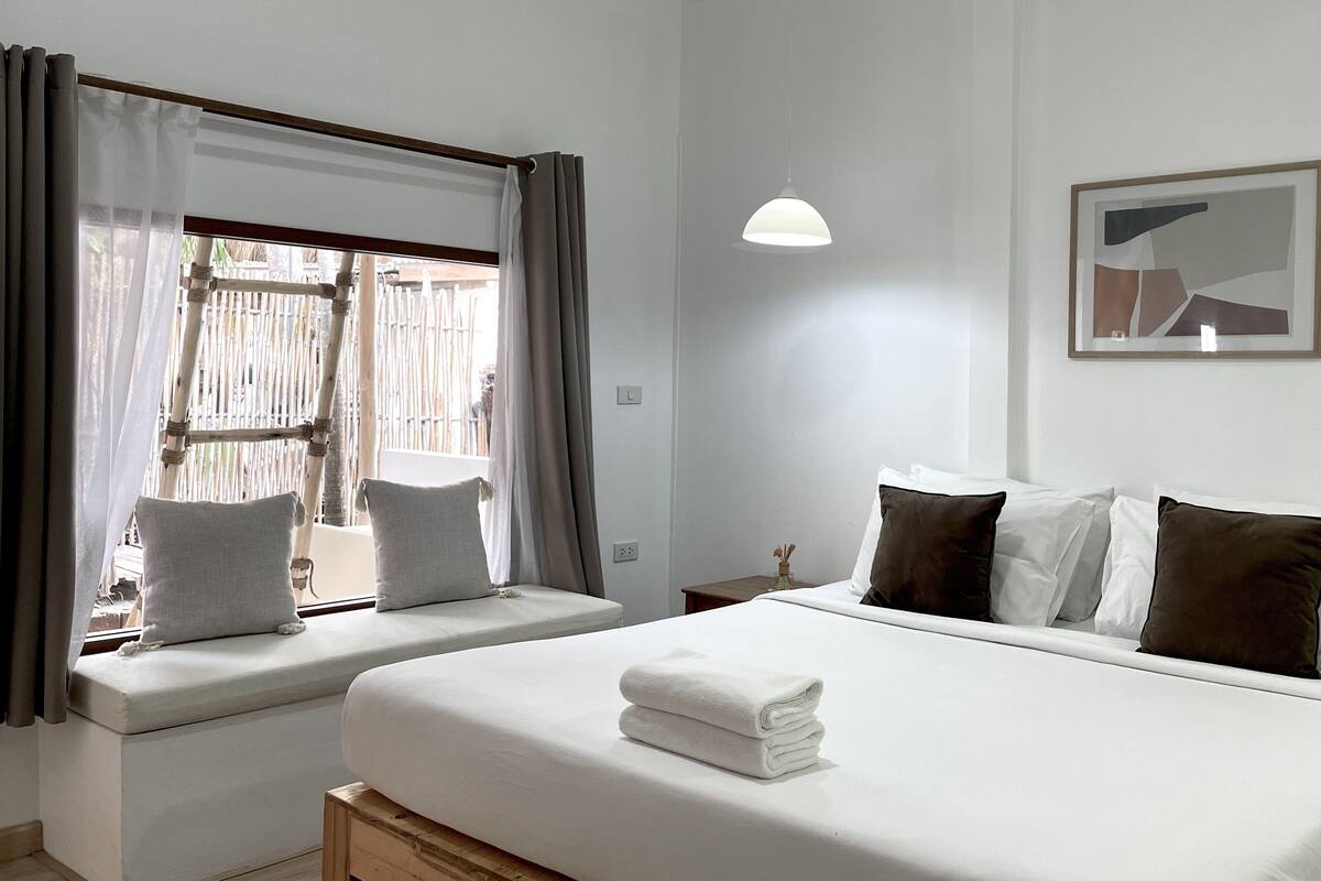 Quarto de hotel com duas camas, uma mesinha de cabeceira, uma luminária e uma janela com cortinas.