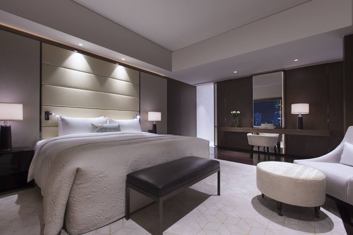 Una habitación de hotel moderna con una cama grande, un banco, un otoman redóondo, un escritorio y una silla.