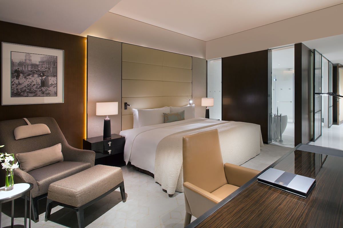 Una habitación de hotel moderna con una cama grande, una zona de estar con una silla, un escritorio y un separador de vidrio que conduce a un baño.