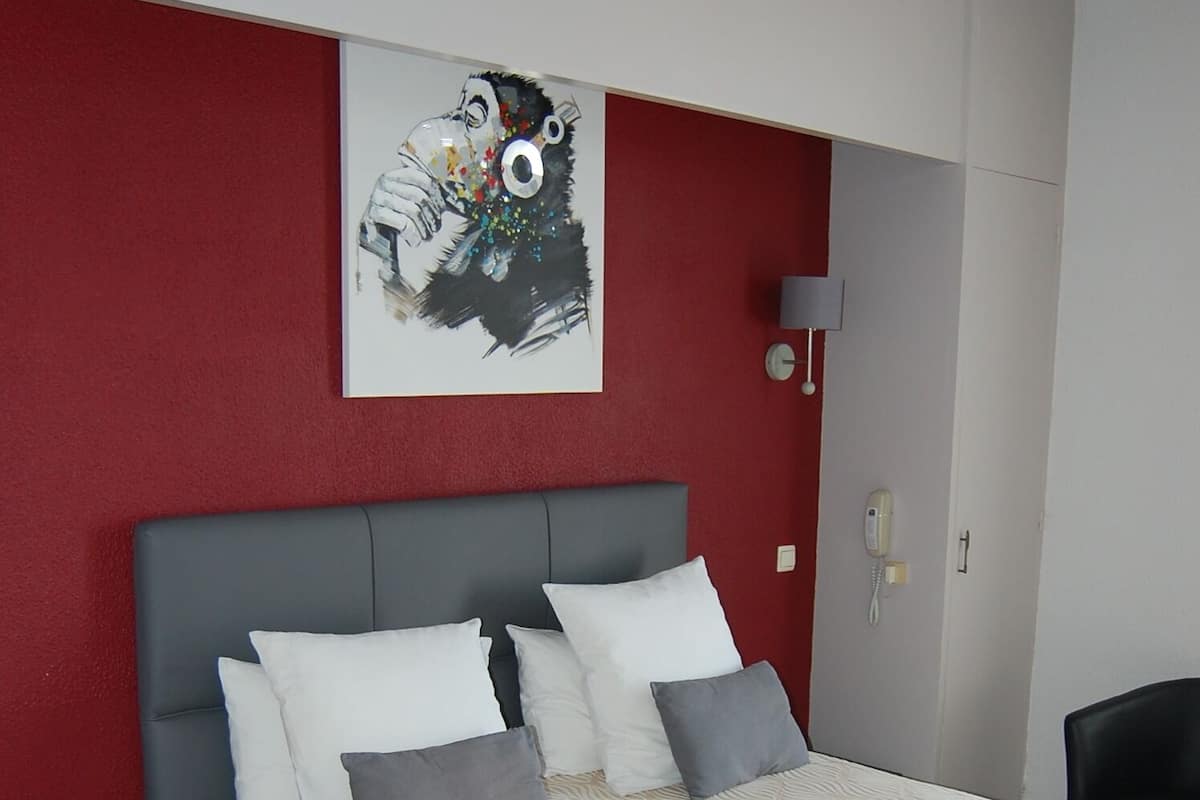 A bedroom with a bed, pillows, a bedside table, a chair, a wall-mounted phone, and a painting.