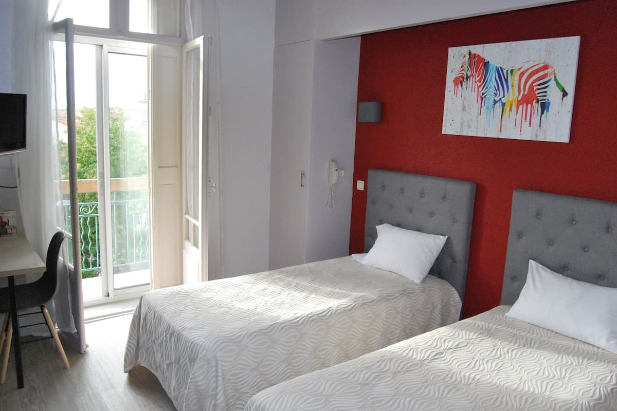 A hotel room with two beds, a desk, and a chair. There is a balcony with a view of greenery.
