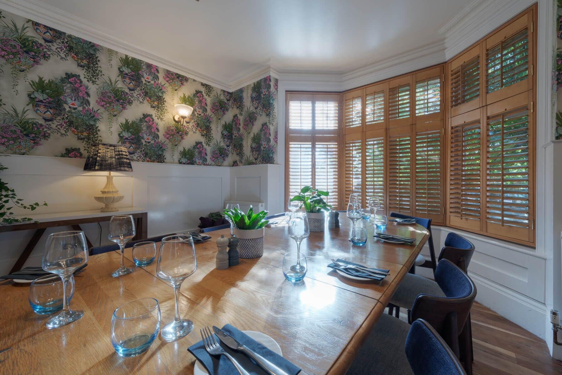 A dining area with a long wooden table set for a meal, surrounded by wooden cabinets with glass doors, and a floral wallpaper design on the walls.