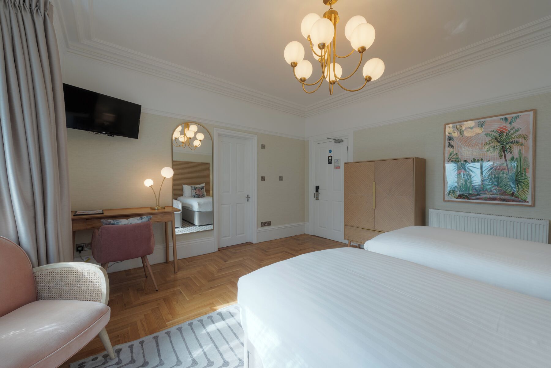 A hotel room with a large bed, a desk with a chair, a mirror, a chandelier, and a painting on the wall.