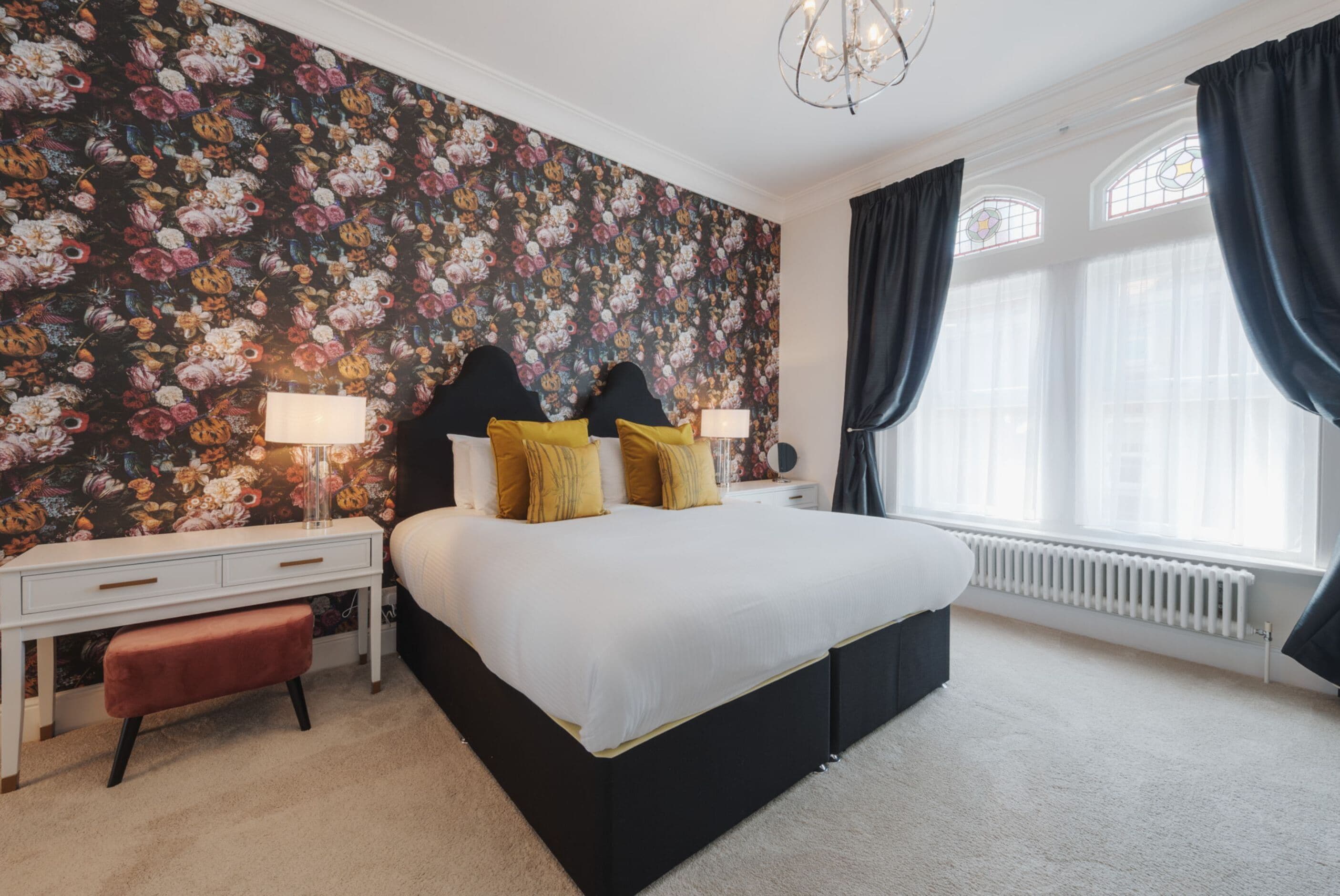 A bedroom with a floral wallpaper, a bed with black headboard, white bedding, and a nightstand with lamps.