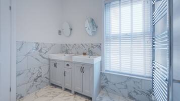 Une salle de bain avec deux lavabos, des carreaux de marbre, une fenêtre avec des stores et un porte-serviettes.