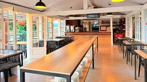Un restaurante con largas mesas de madera y sillas negras, una zona de bar y amplios ventanales que dejan entrar luz natural.