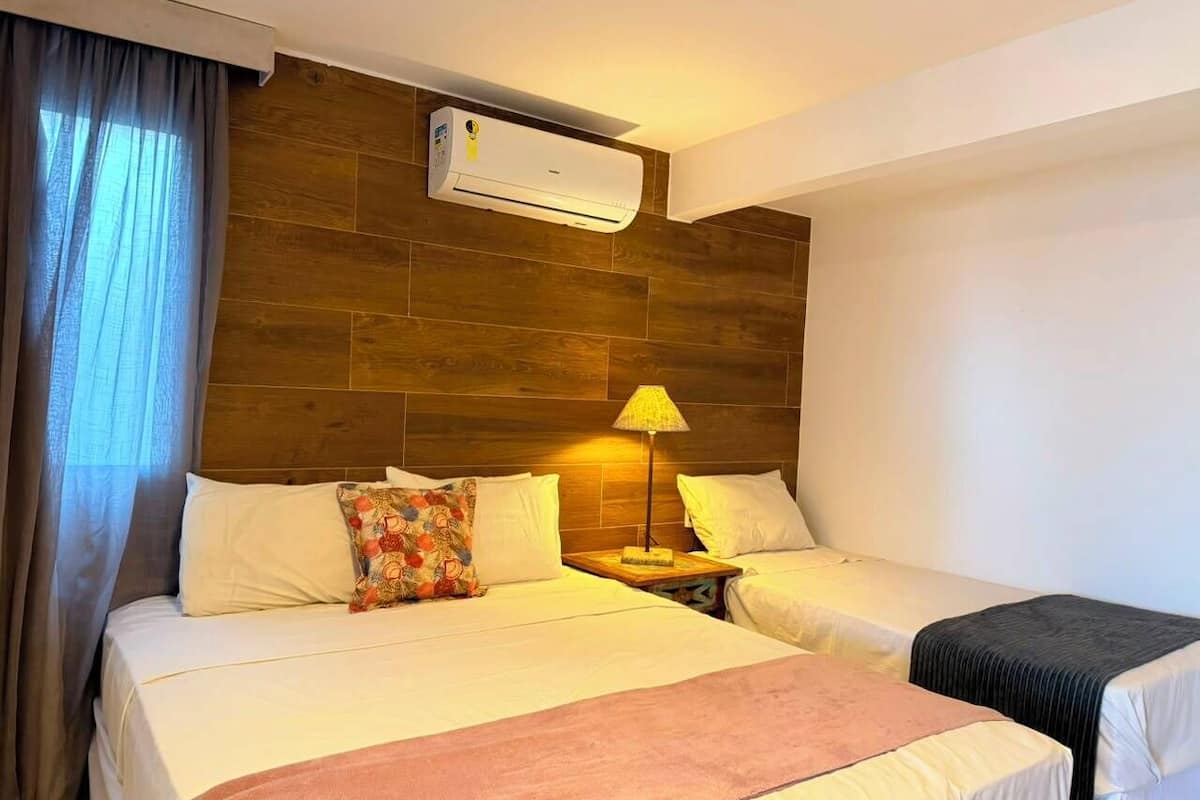 A hotel room with two beds, a nightstand with a lamp, an air conditioning unit, and a window with curtains.