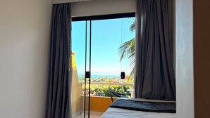 Una habitación con cama, puerta corrediza de vidrio que da a un balcón con vista a vegetación y un paisaje lejano.