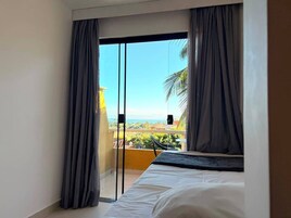 Quarto com cama, porta de correr de vidro para varanda com vista para áreas verdes e paisagem distante.