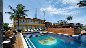 Un área de piscina con sillones de descanso, una terraza de madera y un edificio con múltiples ventanas y balcones.