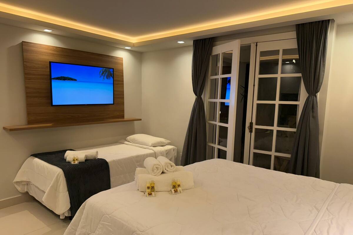 A hotel room with a large bed, two smaller beds, a TV mounted on the wall, and a sliding glass door.