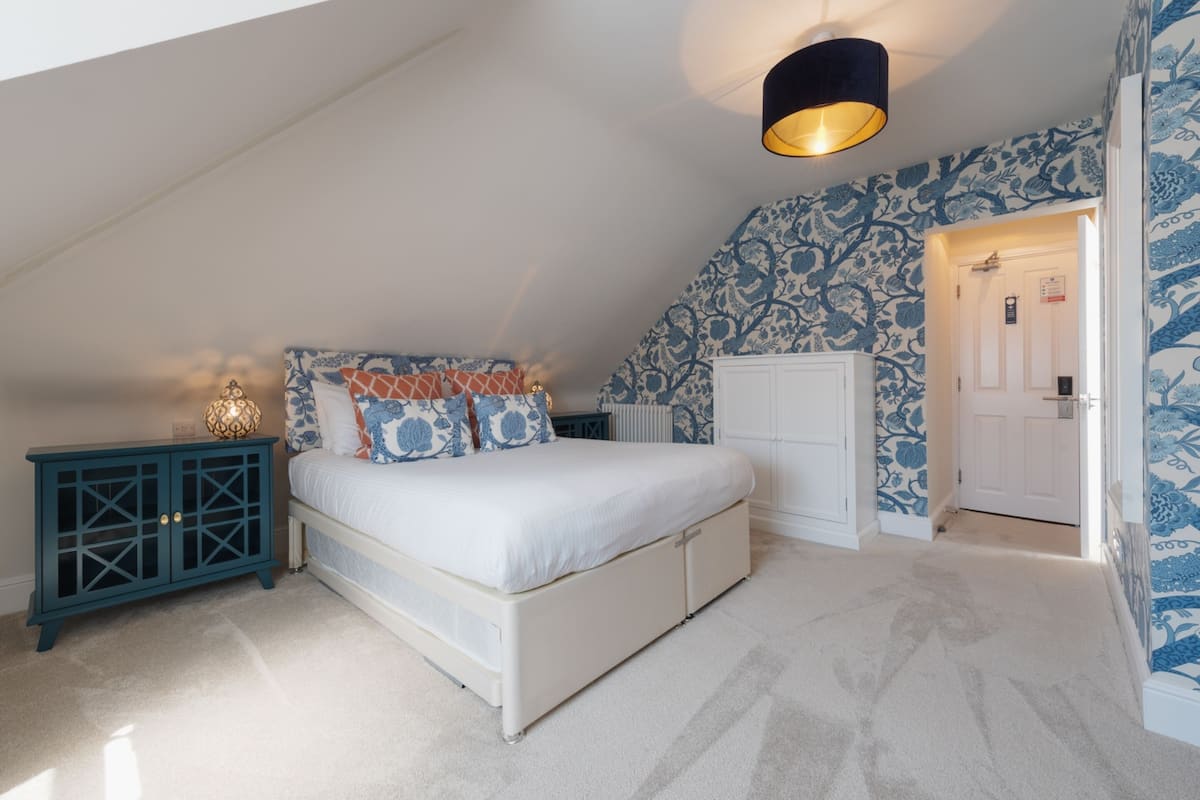 A bedroom with a white bed, blue patterned wallpaper, a teal cabinet, and a ceiling light fixture.