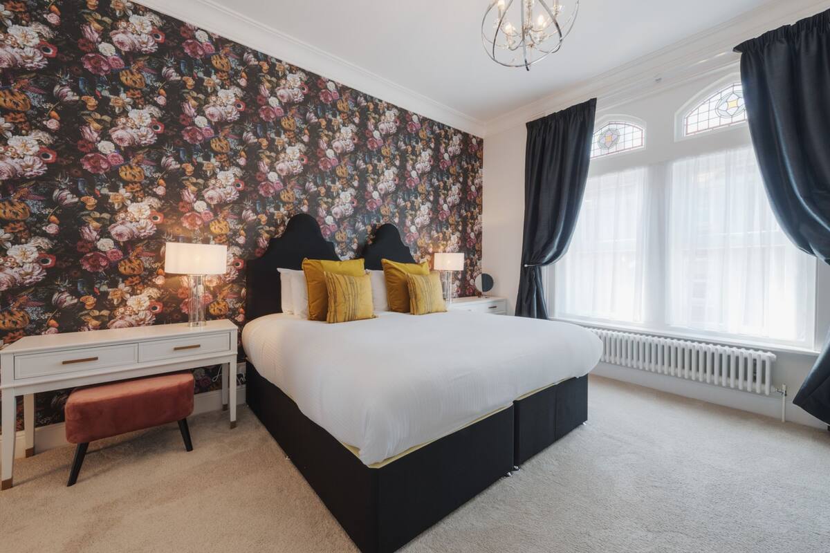 A bedroom with a floral wallpaper, a large bed with black headboard, two bedside tables with lamps, and a window with curtains.