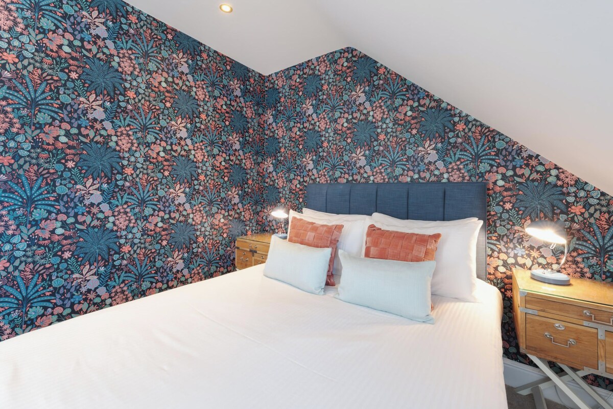 A bedroom with a floral wallpaper, a bed with white bedding, two bedside tables with lamps, and a wooden nightstand.